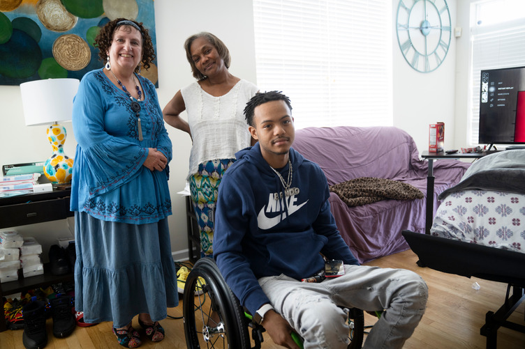 Young man using a wheelchair sits in a living room with two women standing behind him, all smiling toward the camera.