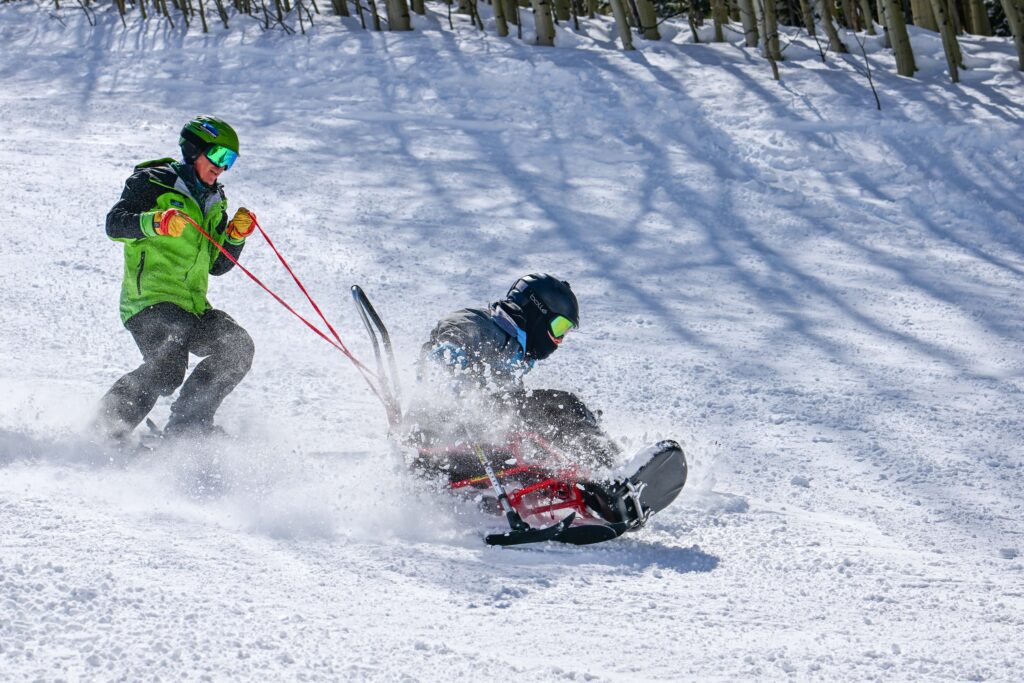 A skier in a bright green jacket assists an adaptive skier using a sit-ski on a snowy slope, guiding them with red tether ropes as they carve through the snow.