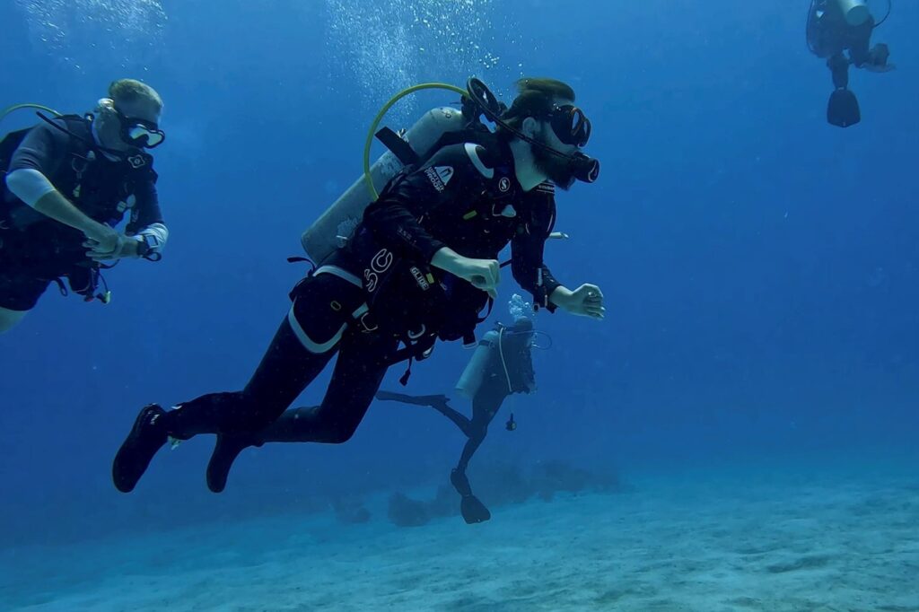 Four scuba divers underwater wearing full diving gear, including wetsuits, masks, regulators, and oxygen tanks, swimming near the sandy ocean floor. The water is clear blue, and small bubbles rise from their breathing equipment.