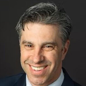 smiling man with short, graying hair, wearing a dark blue suit, light blue shirt and tie