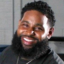 a Black man in his mid-30s with a dark beard and mustache and a smile in his eyes.  He is wearing a black sweatshirt and a silver chain around his neck.