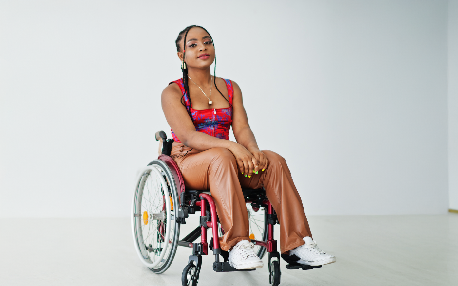 A young woman with braided hair sits confidently in a wheelchair, wearing a colorful top, tan pants, and white sneakers, in a brightly lit studio.